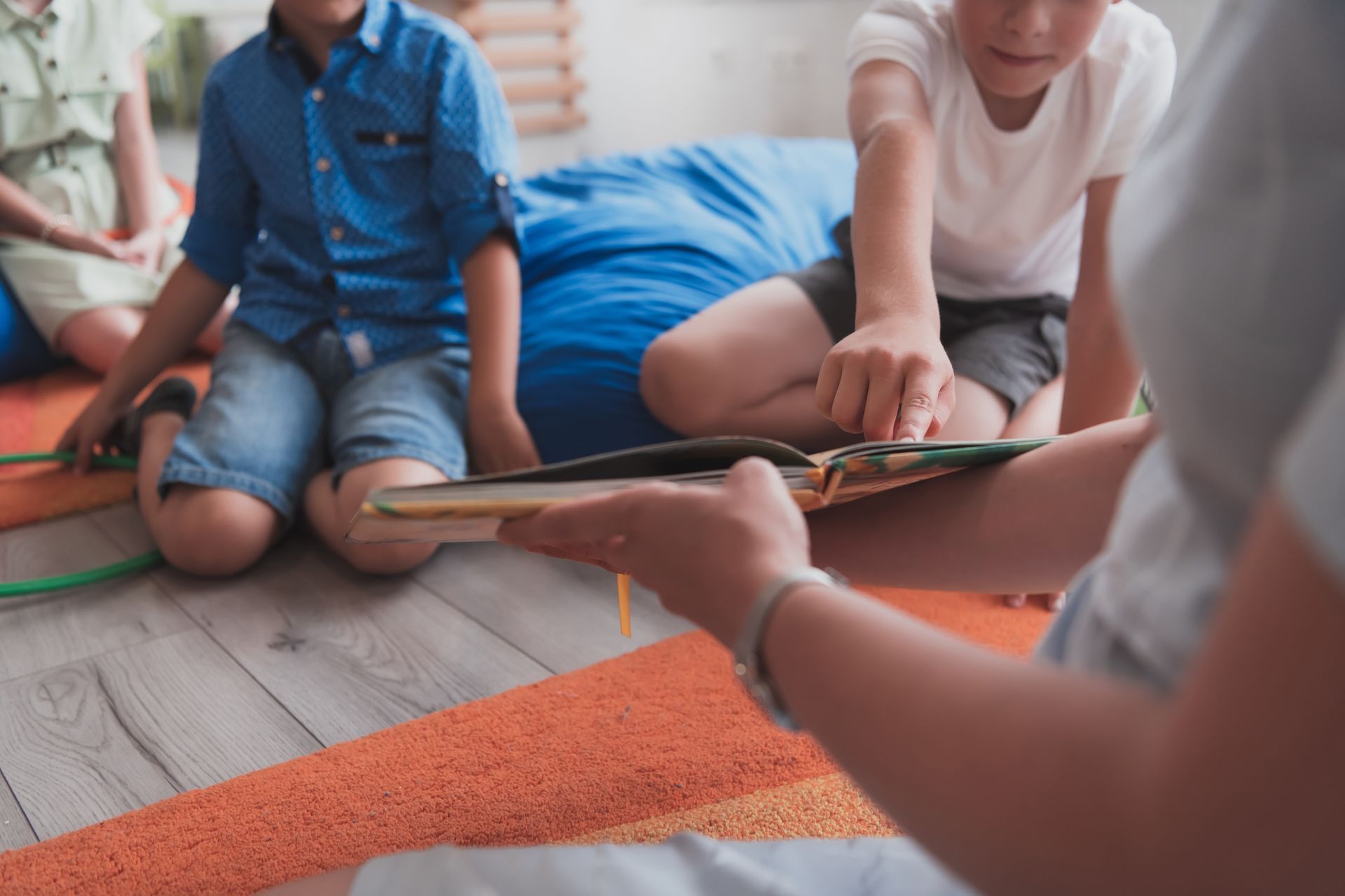 Children sitting on a wooden floor with a book, listening to someone reading on an orange rug.
