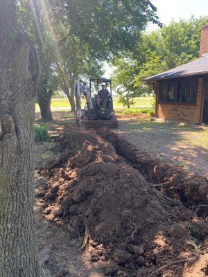 An excavator digging a trench next to a house under a tree.