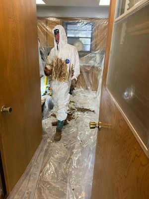 Person in protective suit, splattered with debris, standing in doorway of plastic-sealed room.