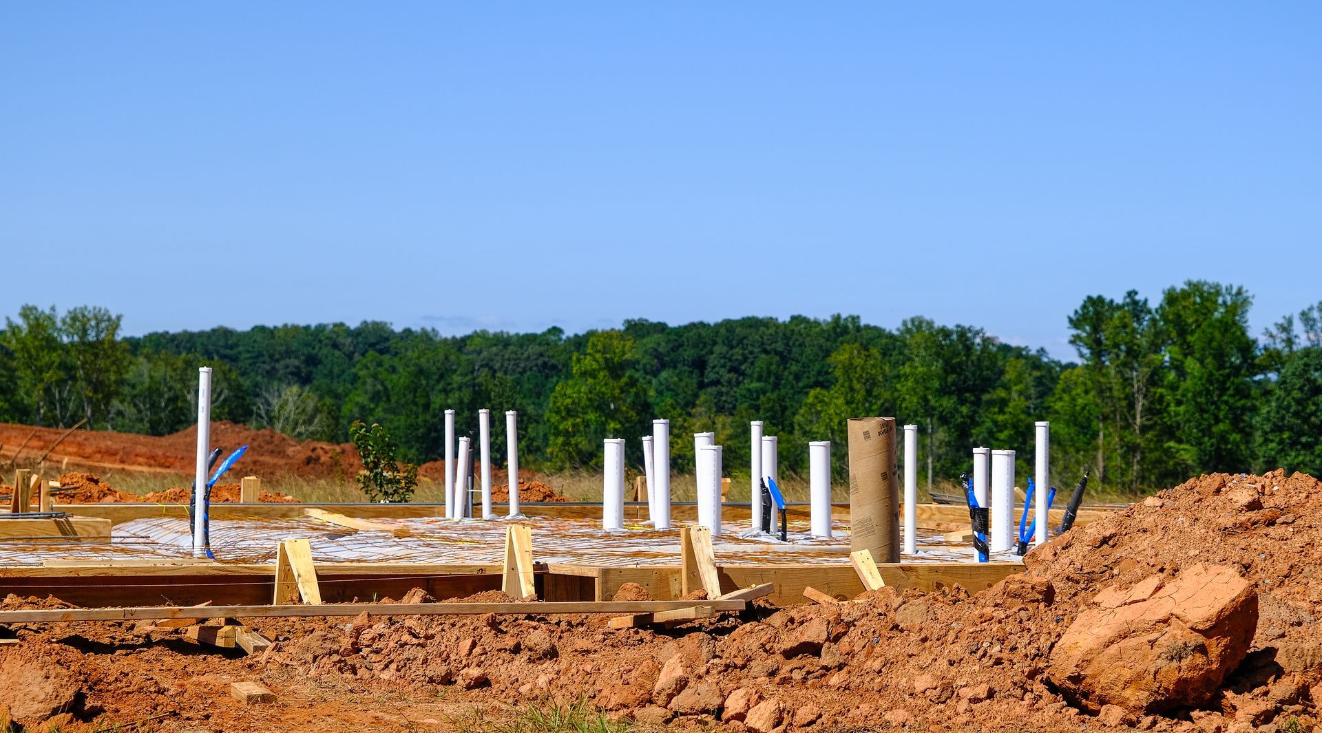 Construction site with foundation and vertical pipes against a backdrop of trees and a blue sky.