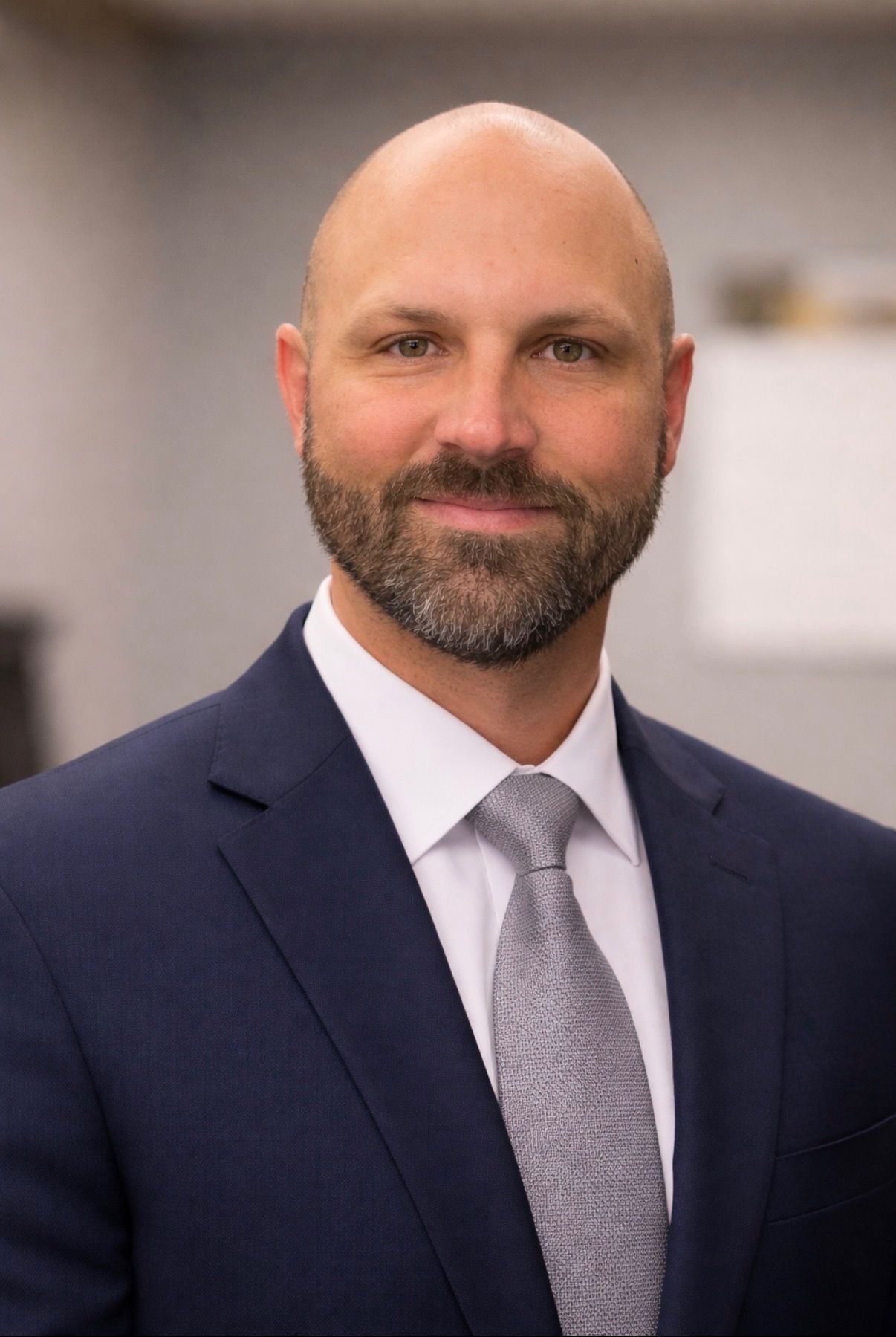 Portrait of a bald, bearded man in a navy suit and gray tie, smiling in an office setting