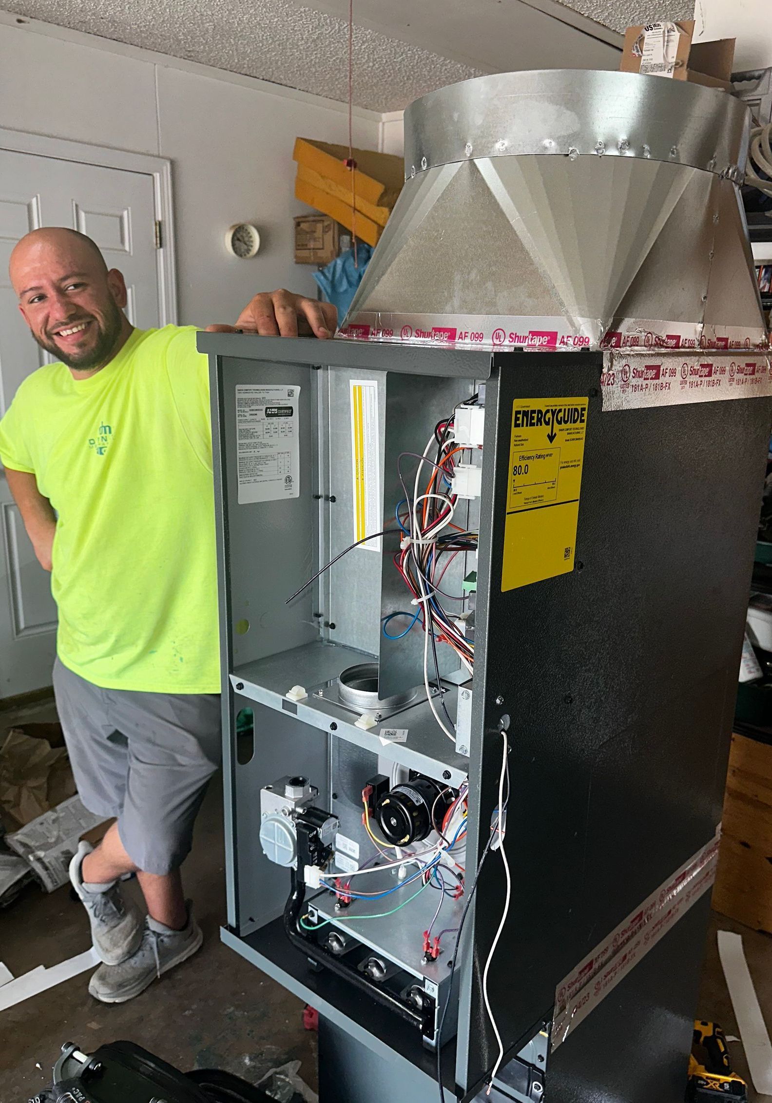 Man in yellow shirt smiles, leans against an open furnace in a garage.
