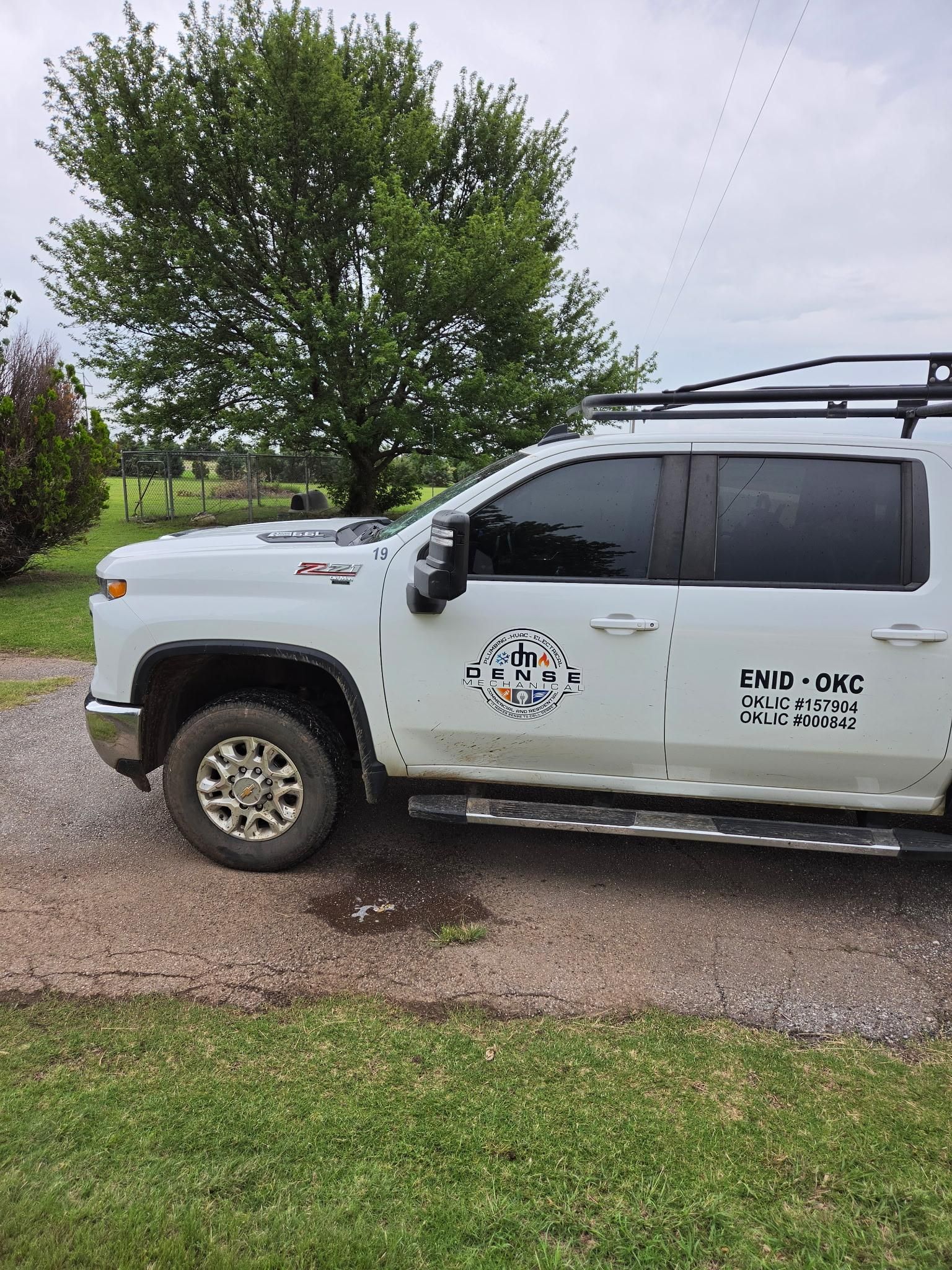 White utility pickup truck parked on gravel beside a green lawn and tree under an overcast sky