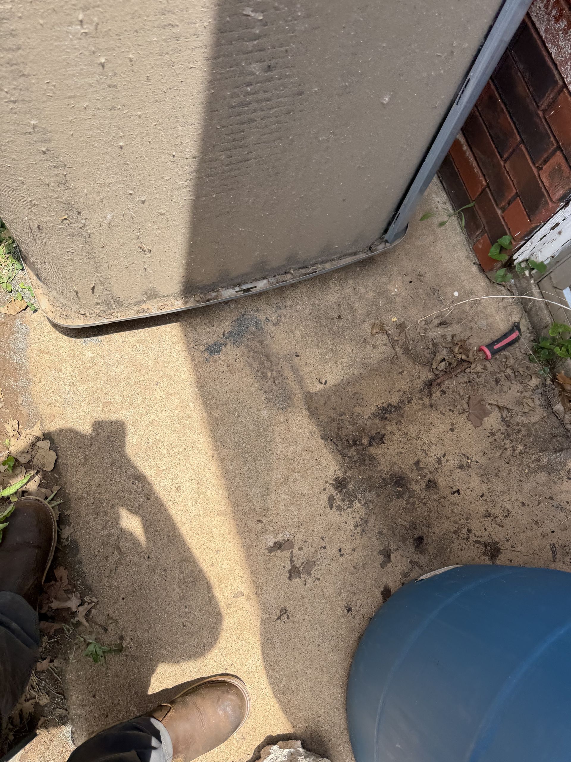 Concrete patio corner with boots, a blue barrel, and dirt near a wall and brick fence.