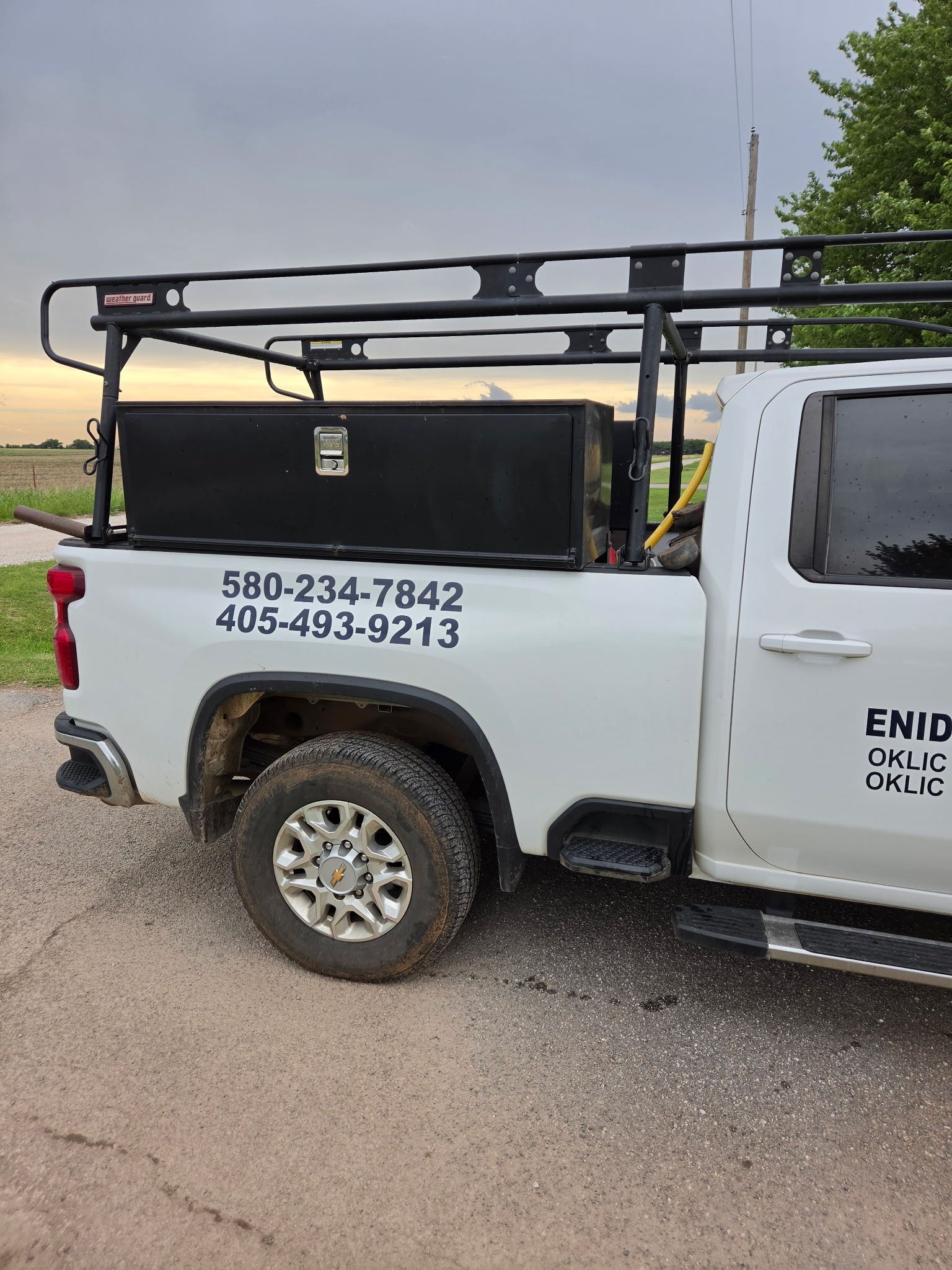 White utility truck with ladder rack and phone numbers on the side, parked on a gravel roadside.