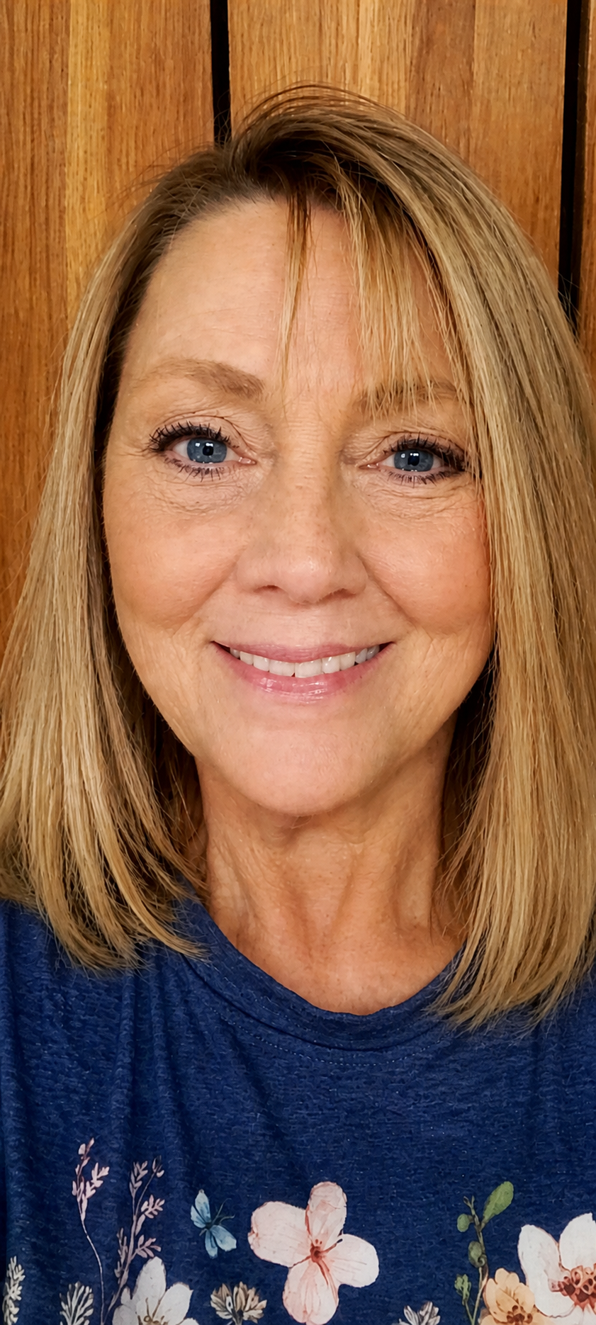 Smiling woman in a blue floral shirt, standing indoors against a wooden background