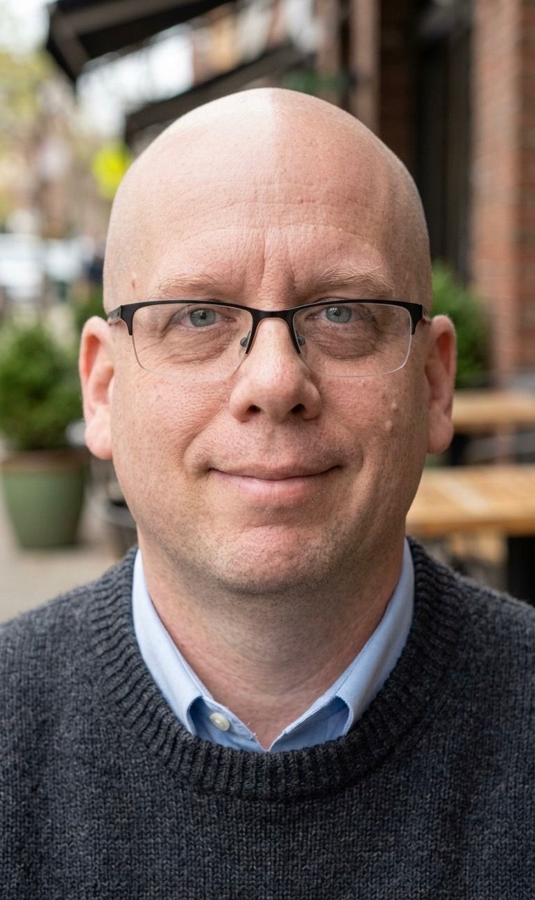 Bald man wearing glasses and a dark sweater, seated outdoors with a blurred café background