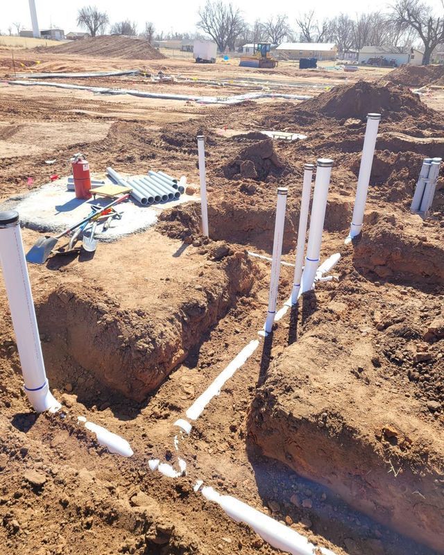 Construction site: PVC pipes in trenches, tools, red tank, exposed soil, bright sunlight.