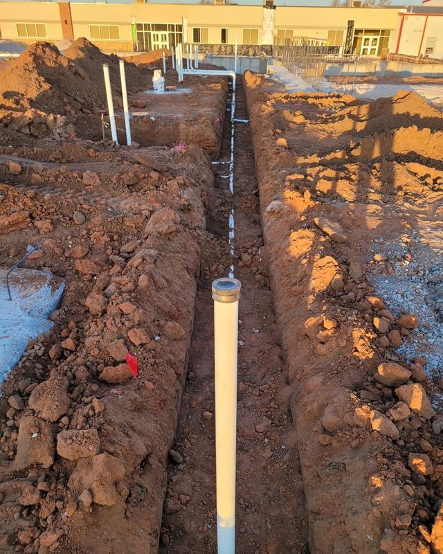 Trench with white pipes, likely for plumbing, running towards a building under construction.