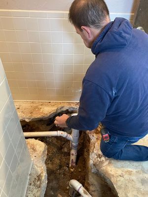 Plumber working on pipes in a bathroom. He is kneeling near the exposed pipes in the floor with a blue sweatshirt on.