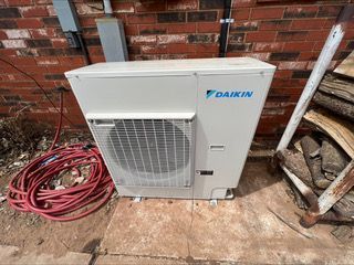 Daikin heat pump unit outdoors against a brick wall, with a red hose and firewood nearby.