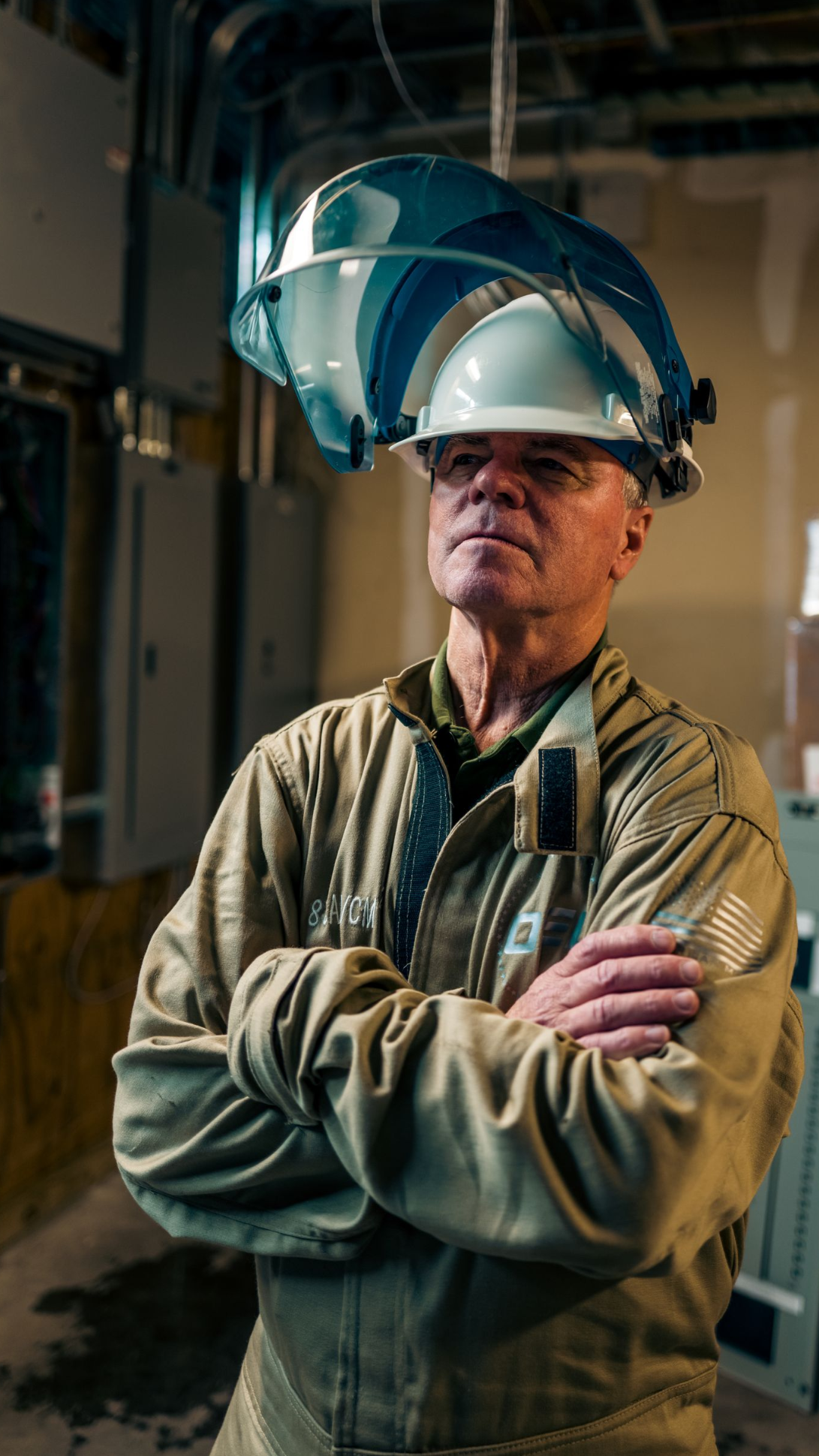 Worker in a hard hat stands with arms crossed in an industrial facility, wearing a raised face shield.