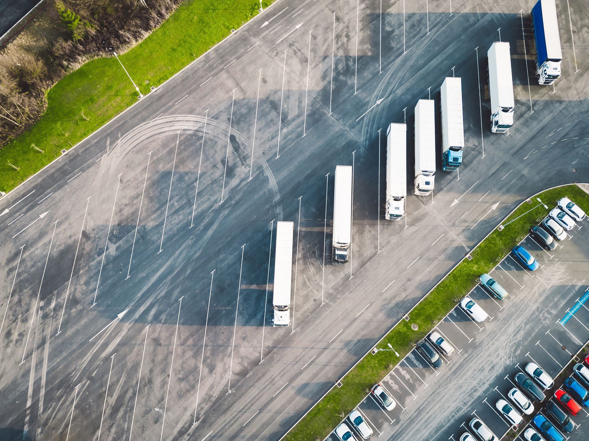 Aerial view of white semi-trucks lined up in a parking lot next to parked cars.