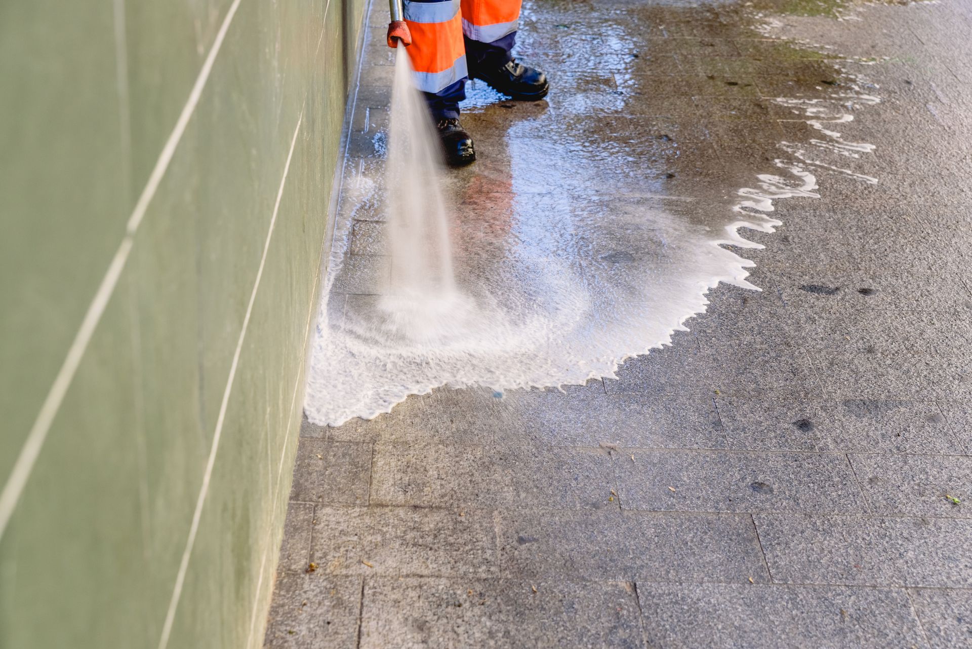 Worker pressure-washing a wet sidewalk beside a green wall, with water spraying onto the pavement