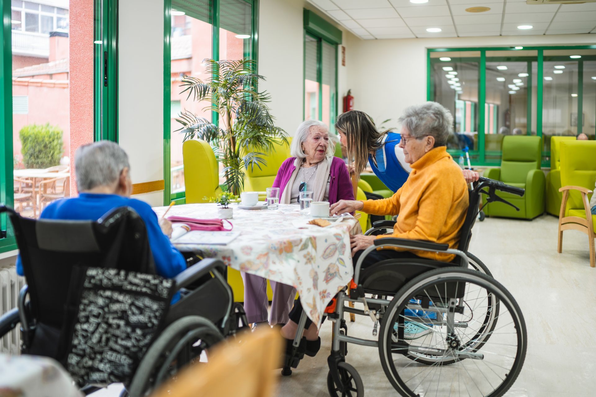 People in wheelchairs socializing around a table in a bright care home lounge