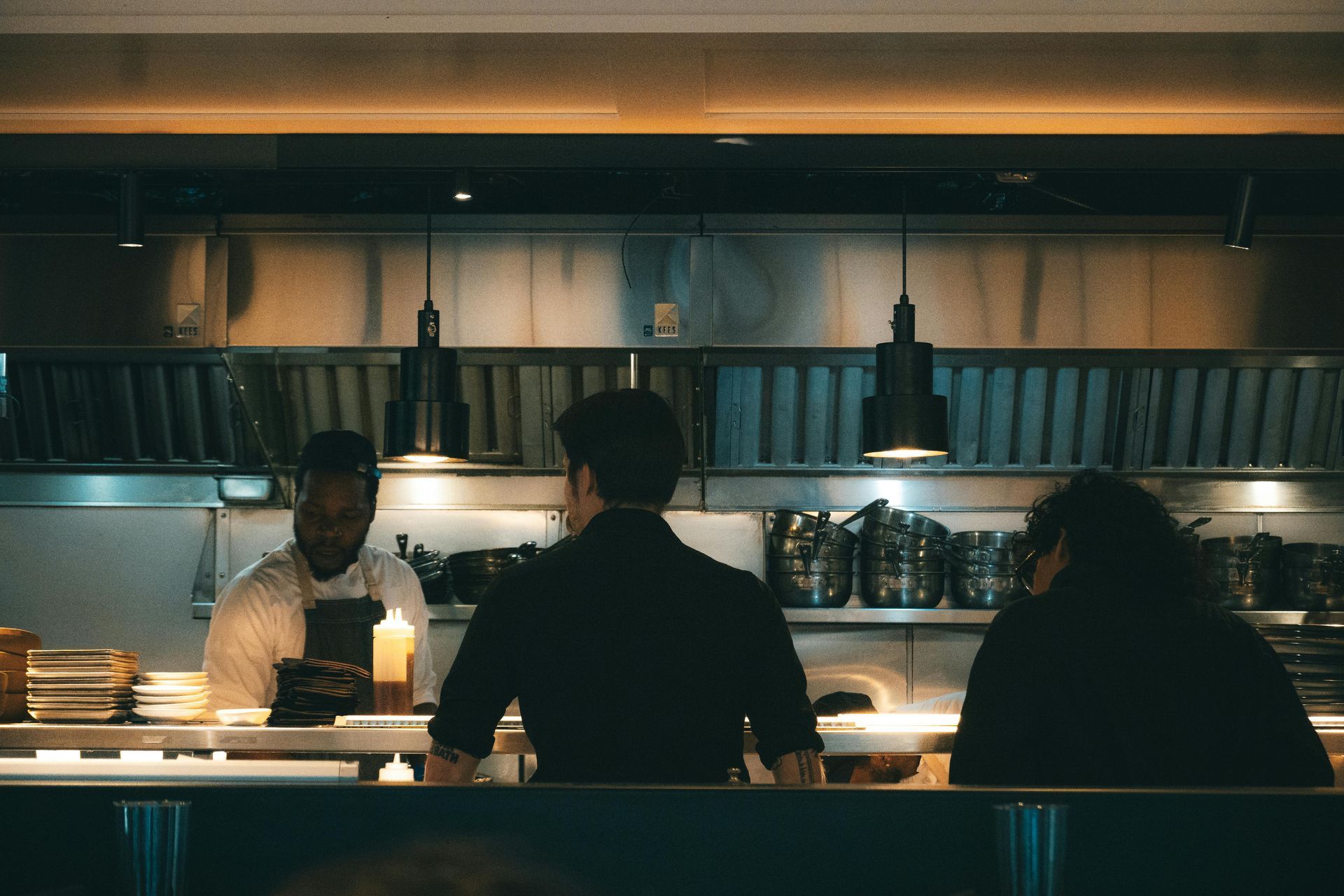 Three people sit at a warmly lit commercial kitchen counter with hanging pendant lights and shelves of cups behind them.