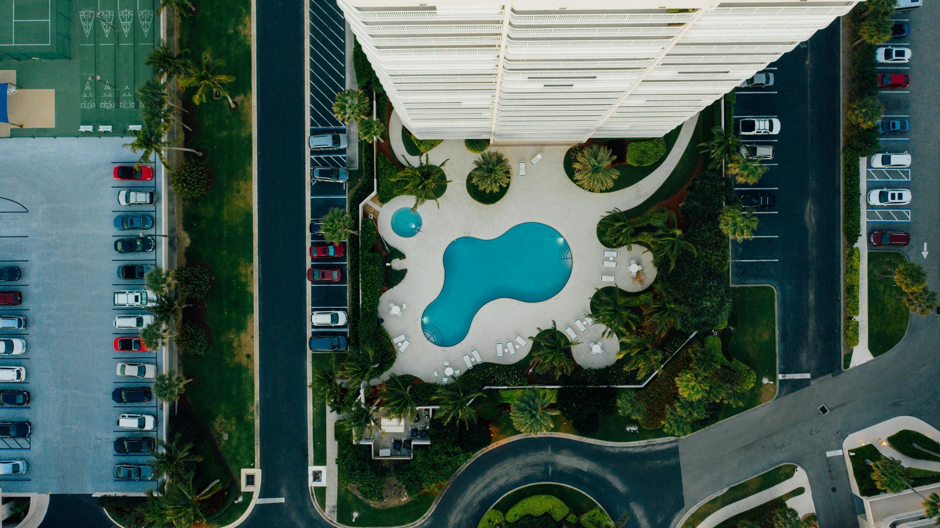 Aerial view of a turquoise pool in a landscaped courtyard beside a building and parking lot.