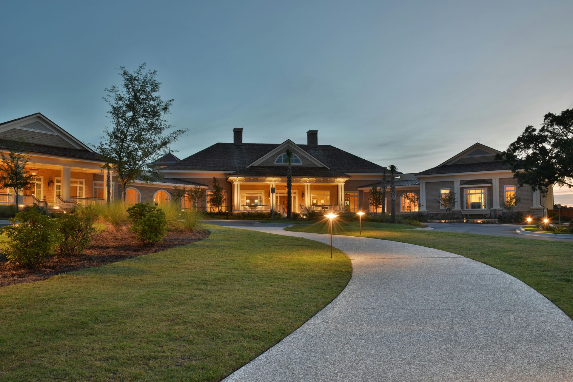 Curved gravel path leading to a lit single-story house at dusk, with manicured lawn and trees