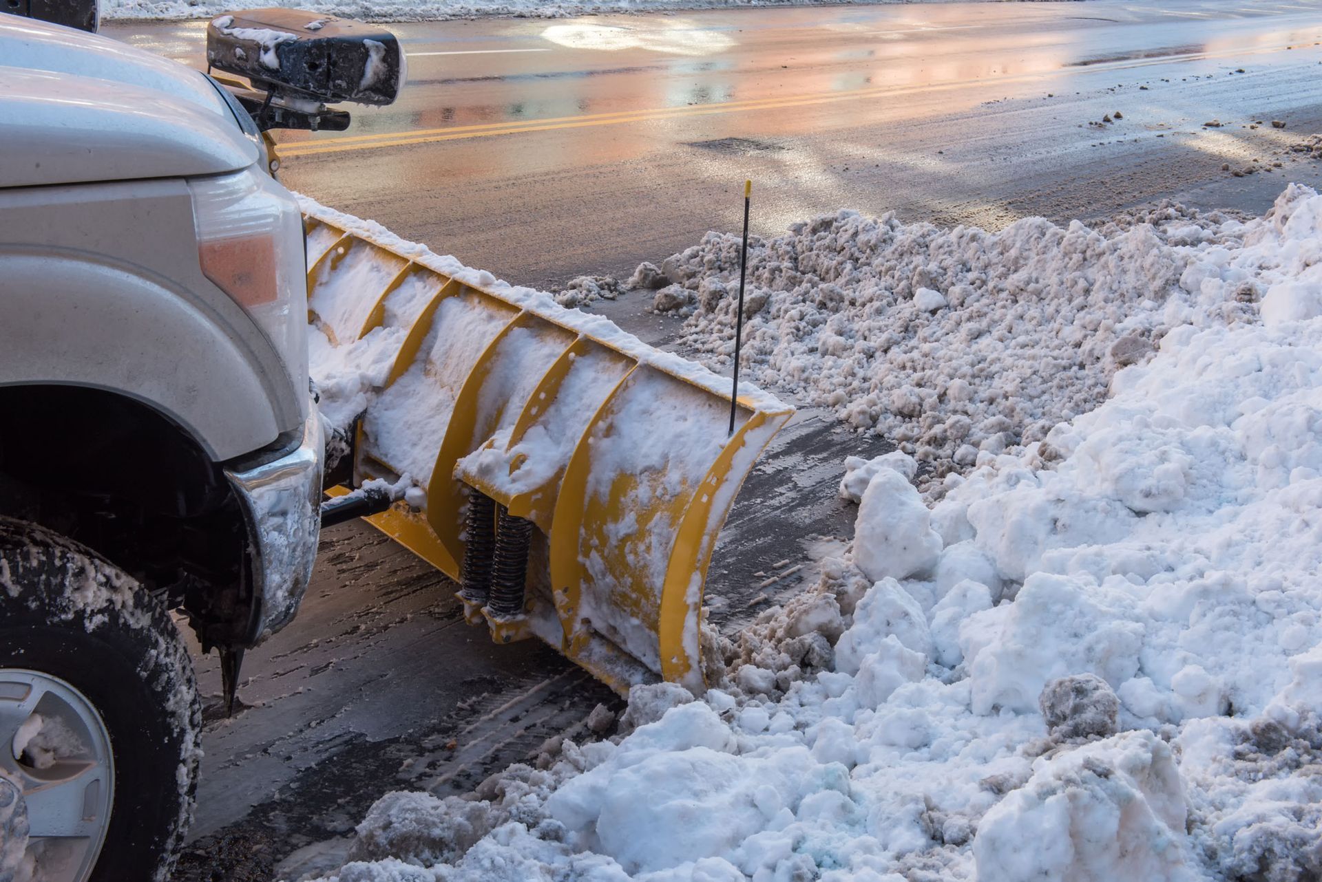 Truck with a yellow snowplow clearing a snowy street at dusk