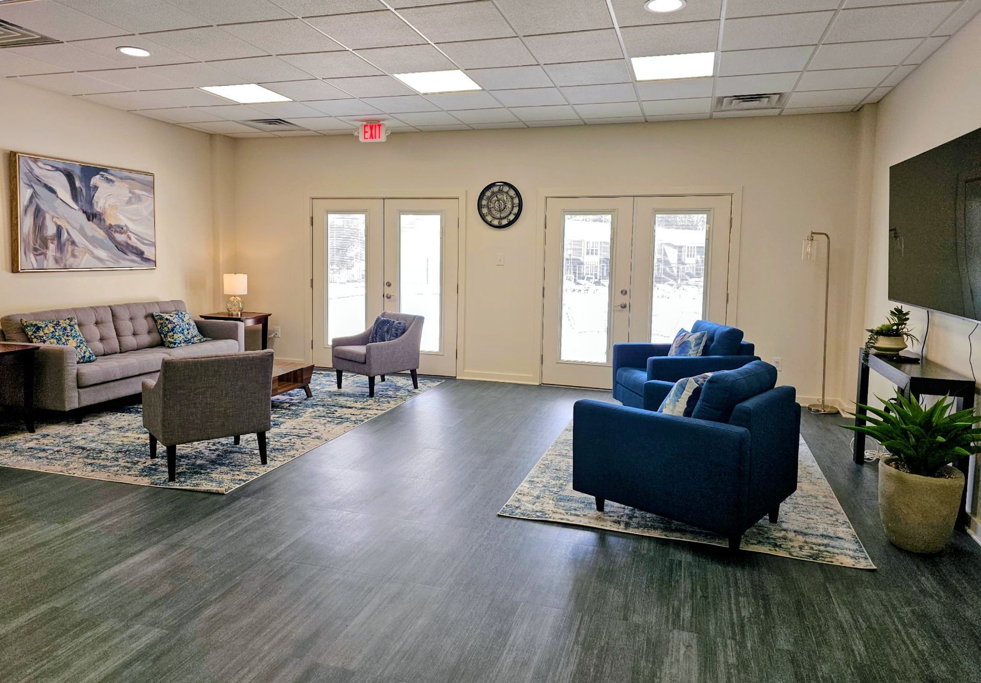 Bright lobby with blue chairs, couch, tables, potted plants, and glass doors under a clock