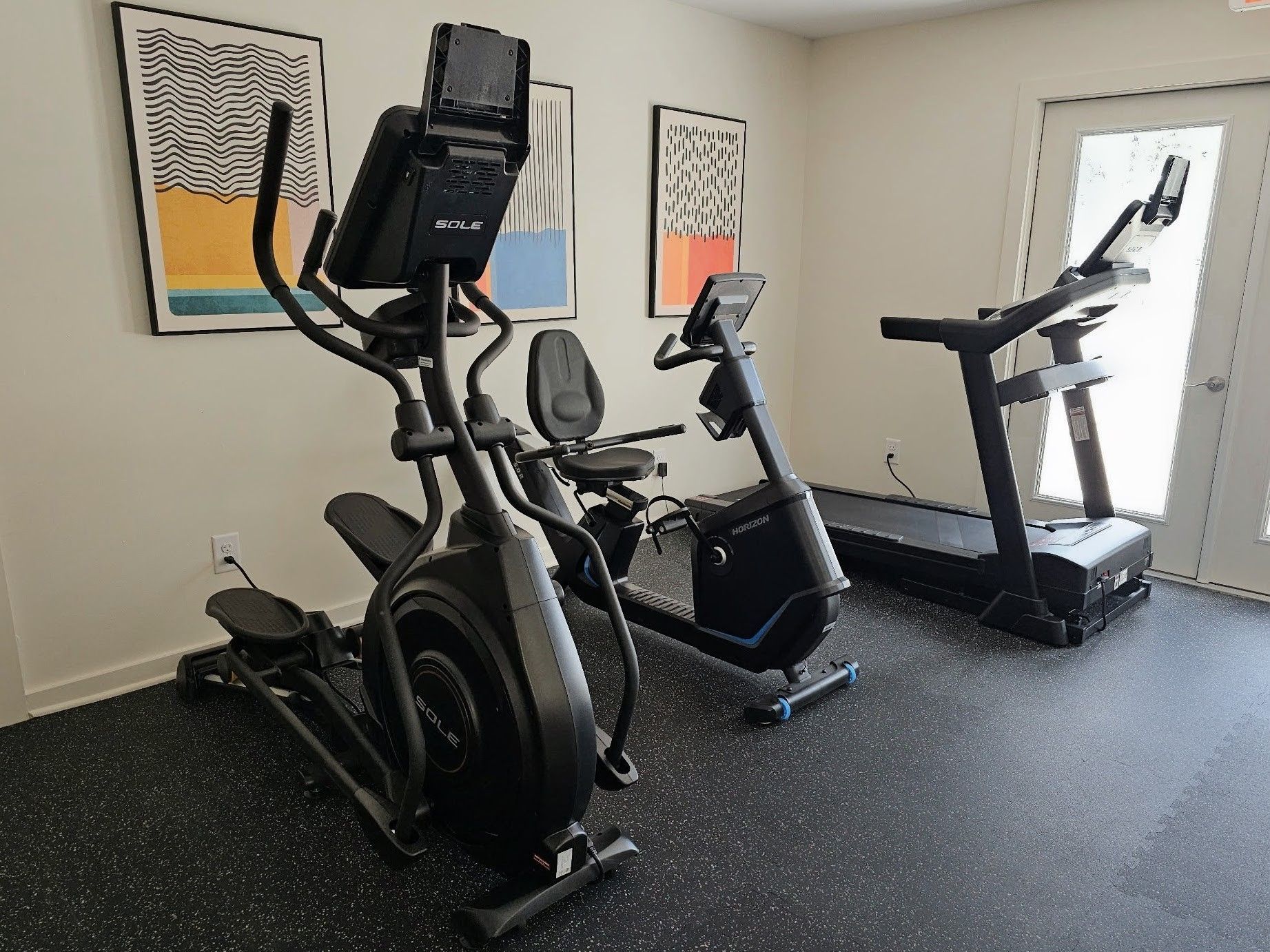 Small gym with an elliptical, stationary bike, and treadmill on rubber flooring.