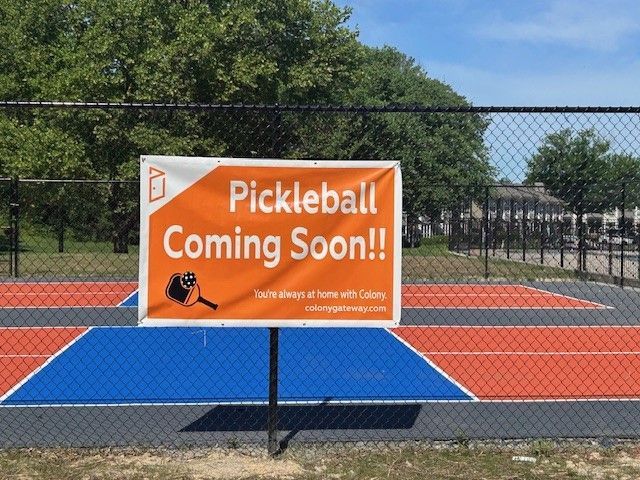 Orange sign on a tennis court reads “Pickleball Coming Soon!!” with a blue temporary court below.