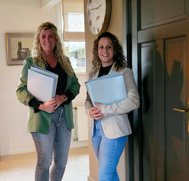 Twee vrouwen in zakelijke kleding, met mappen in hun handen, staan glimlachend in een deuropening van een kantoor.
