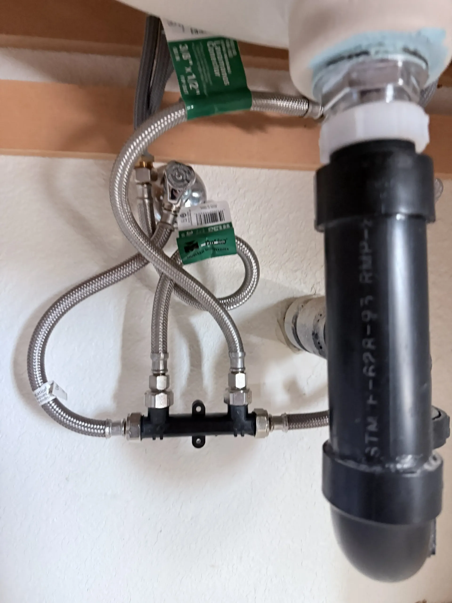 Under-sink plumbing with flexible metal hoses, green labels, and a black drain pipe assembly
