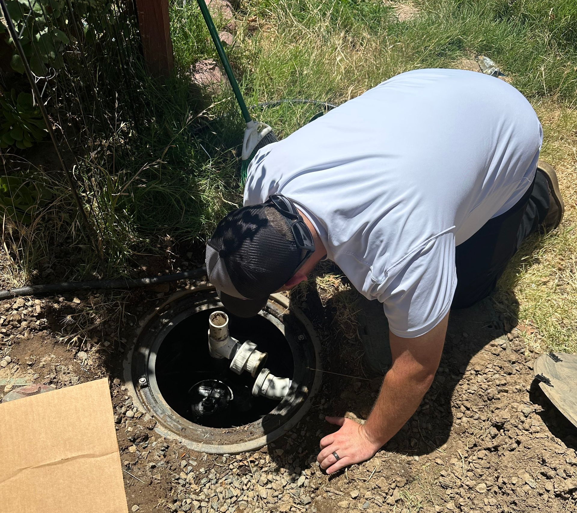 Person bends over an open utility access hole outdoors, inspecting pipes inside.