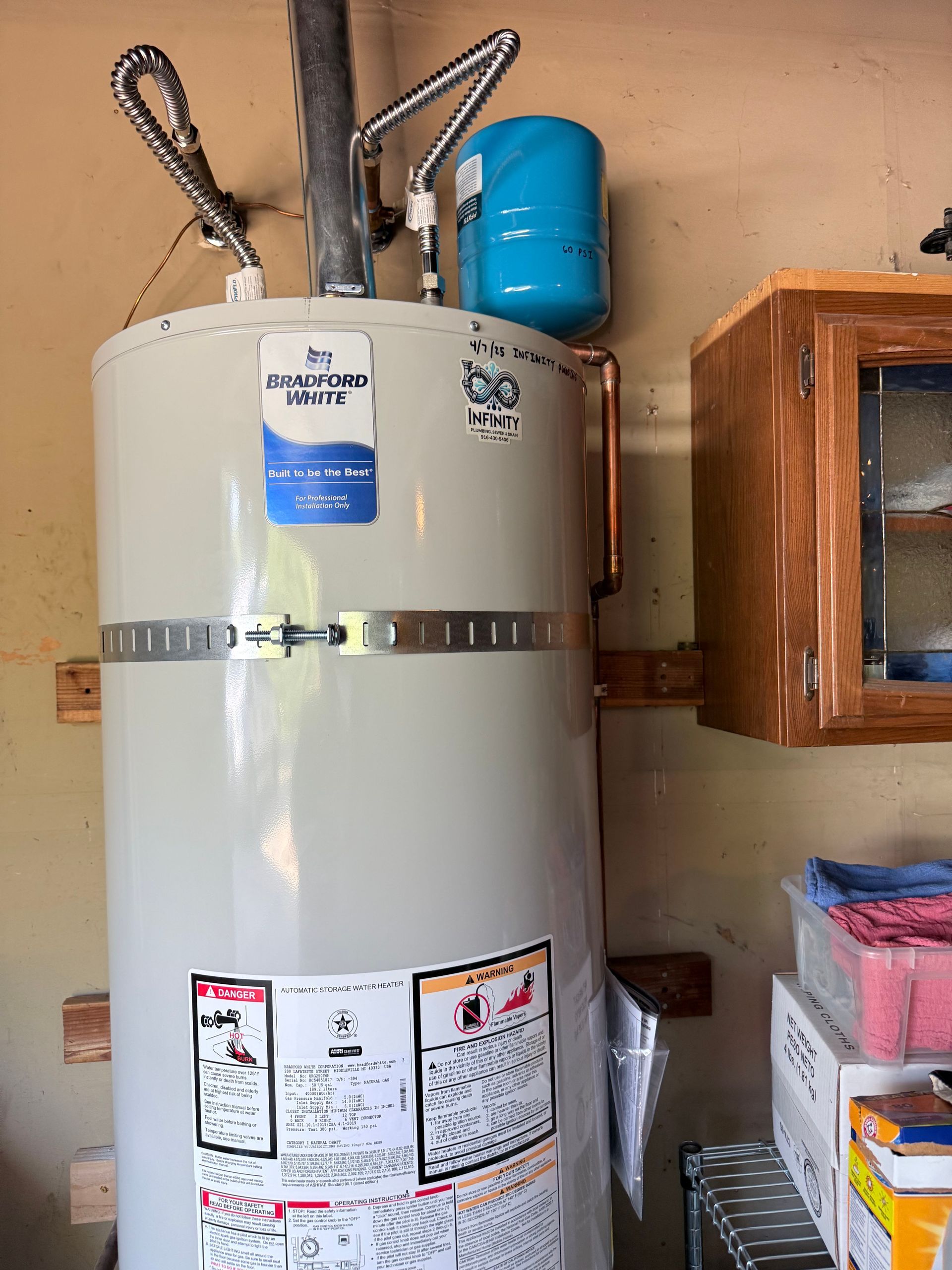 Water heater tank with blue expansion tank and pipes in a utility room, next to shelving.