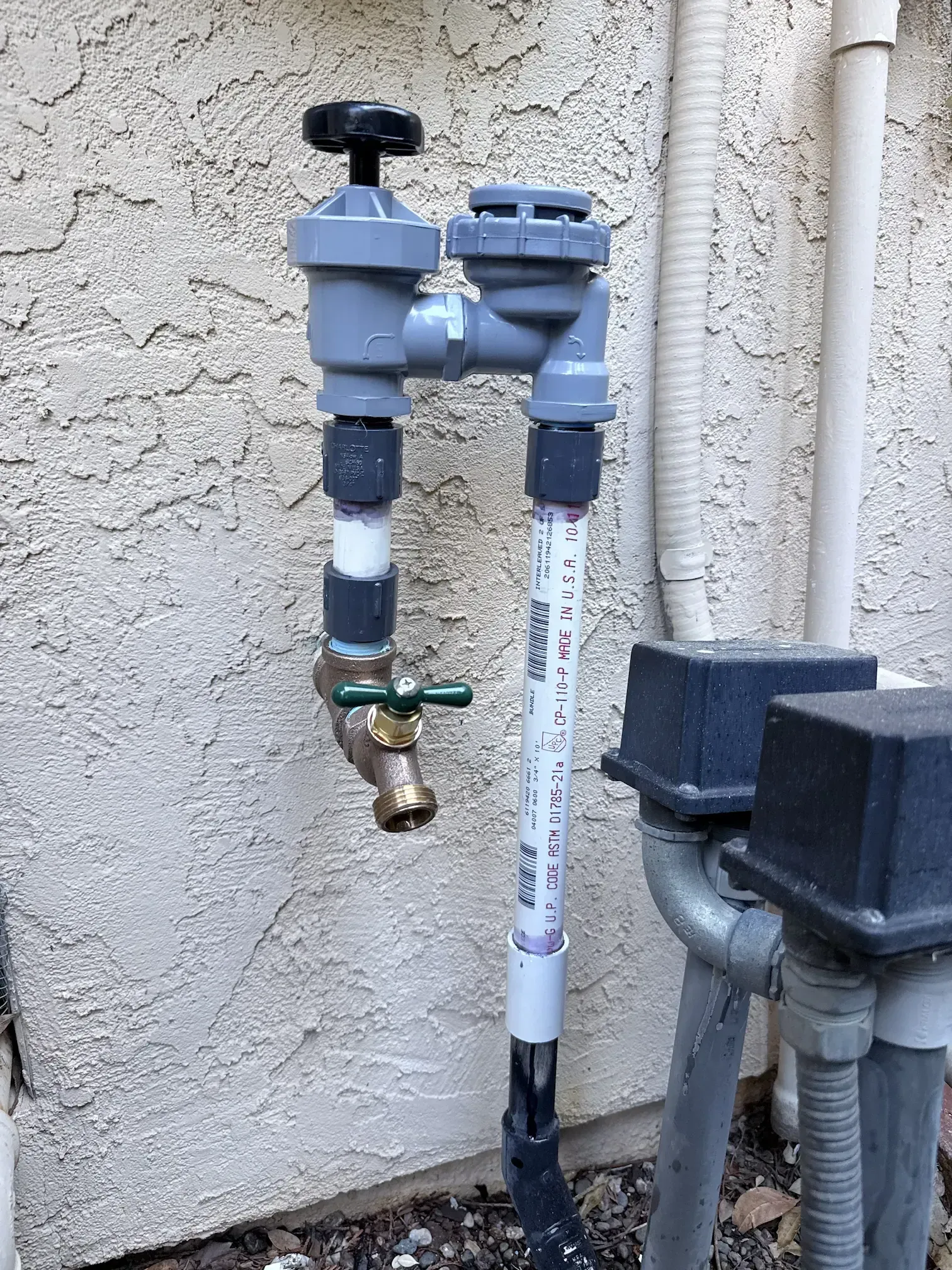Outdoor water meter and backflow valve assembly mounted on a stucco wall, with white PVC pipes and brass faucet.