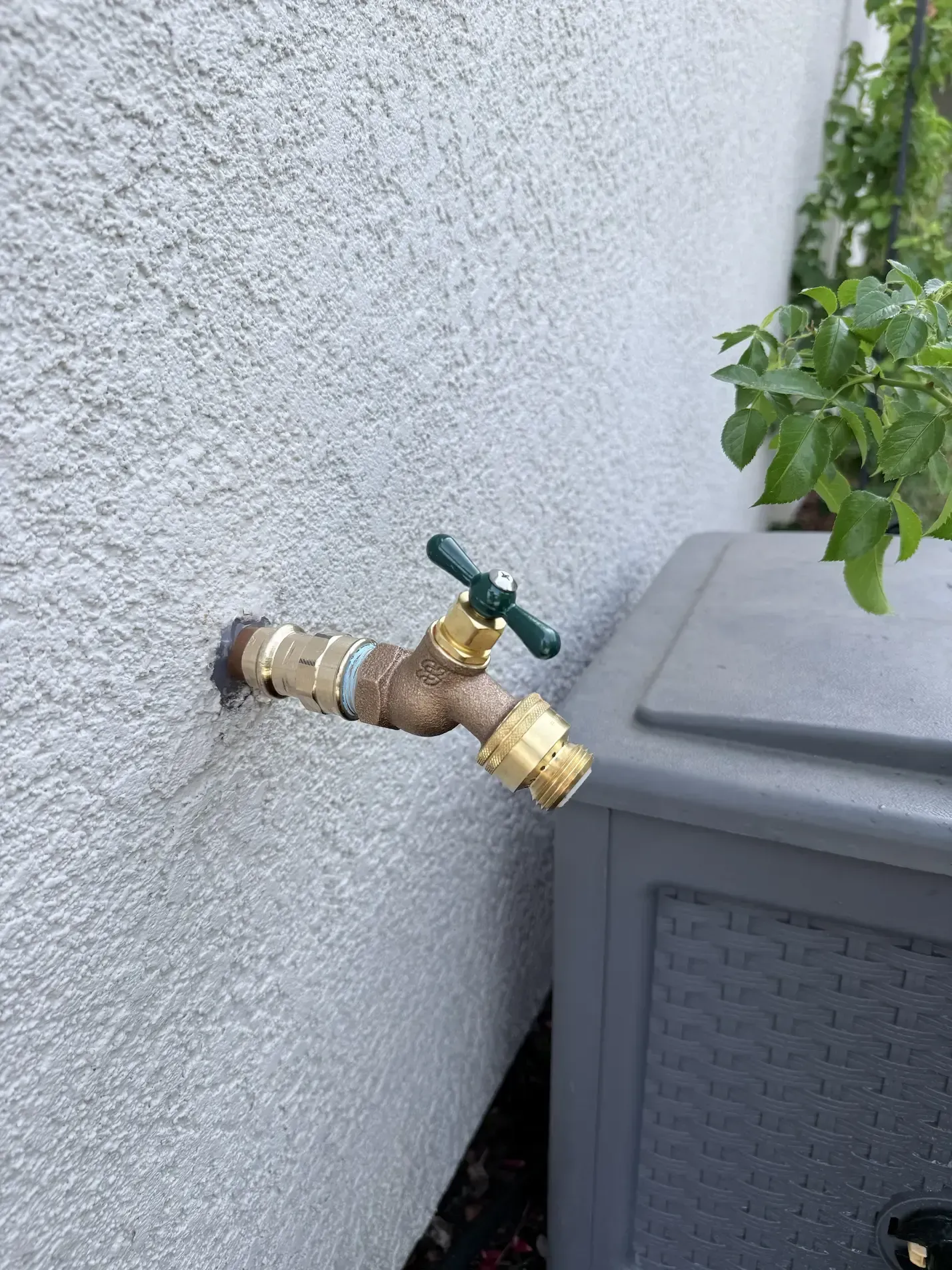 Outdoor wall faucet with green handle mounted beside a gray utility unit
