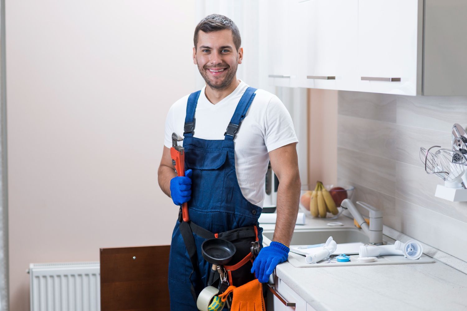 Cleaner in blue overalls holding cleaning tools in a kitchen, smiling beside a counter and cabinets