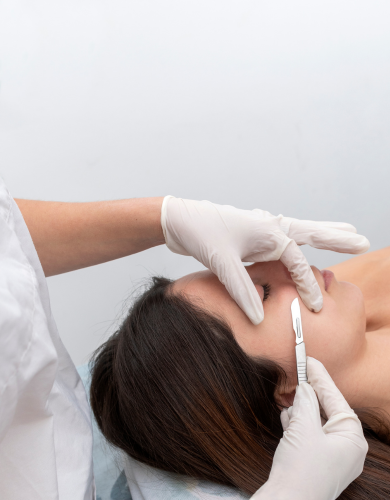 Person receiving a dermaplaning treatment with gloved hands and a scalpel on their face in a white room.