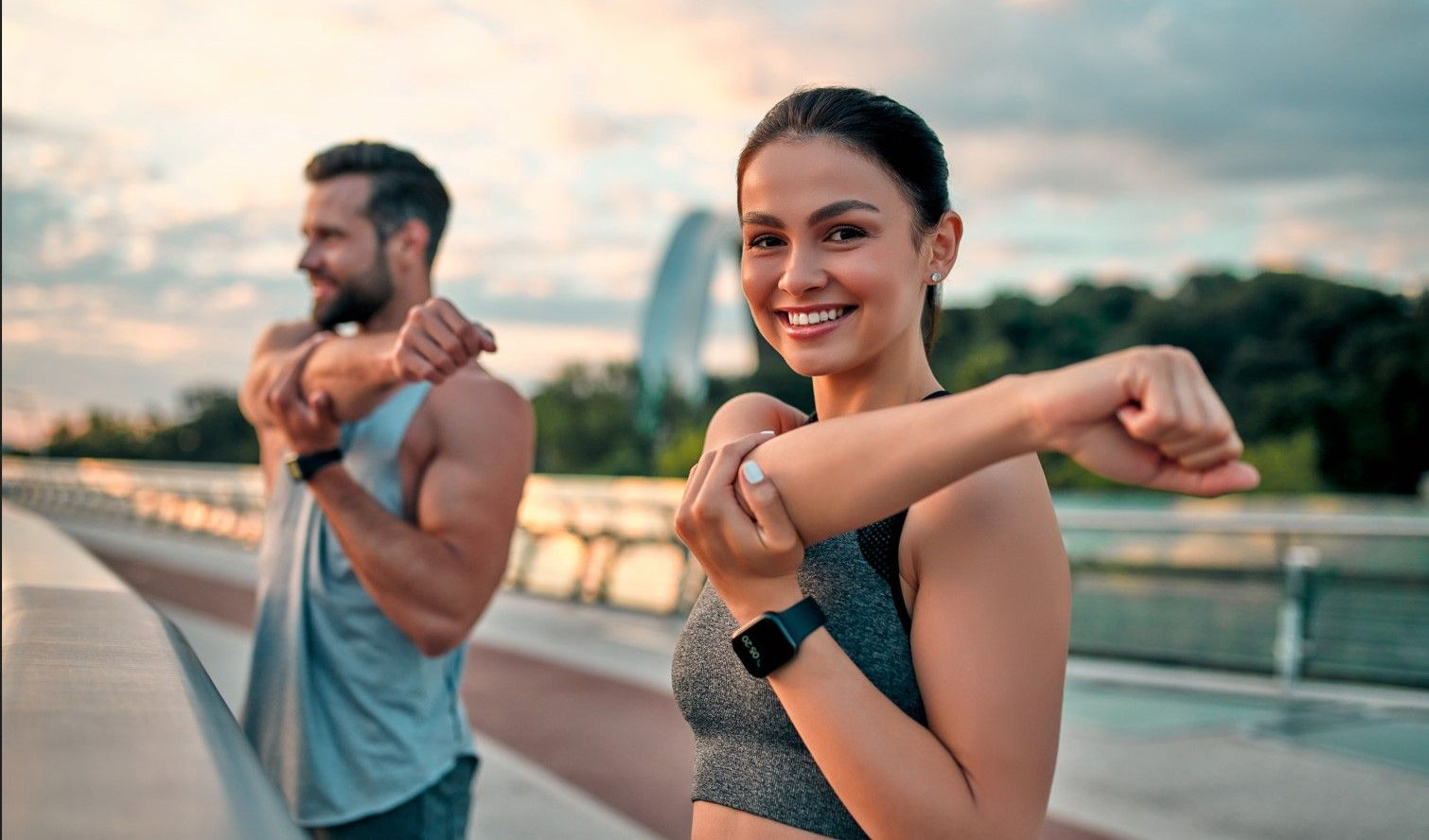 Woman and man stretching arms outdoors, smiling, with cityscape background.