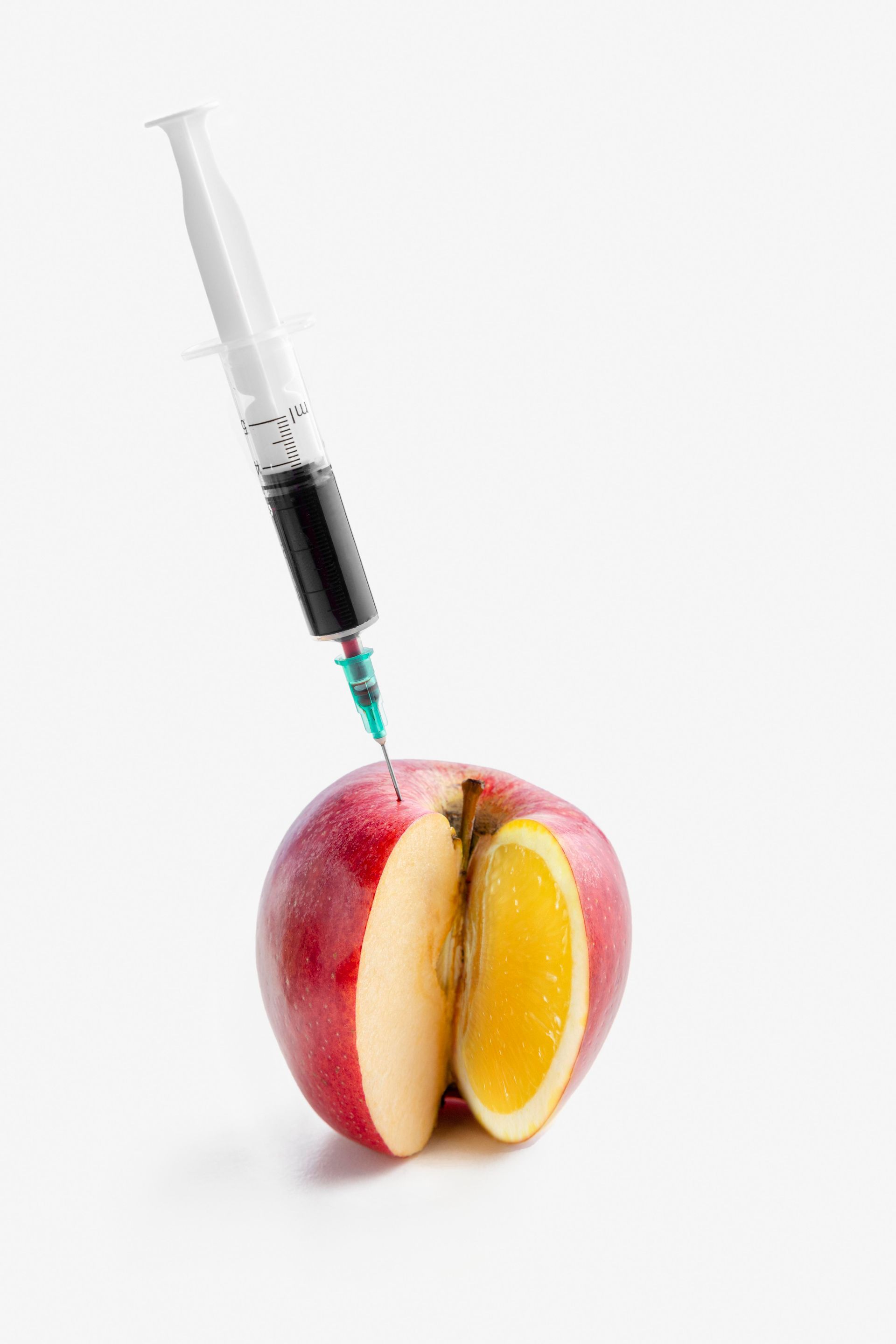 An apple sliced to reveal an orange segment, injected with a syringe of dark liquid against a white background.