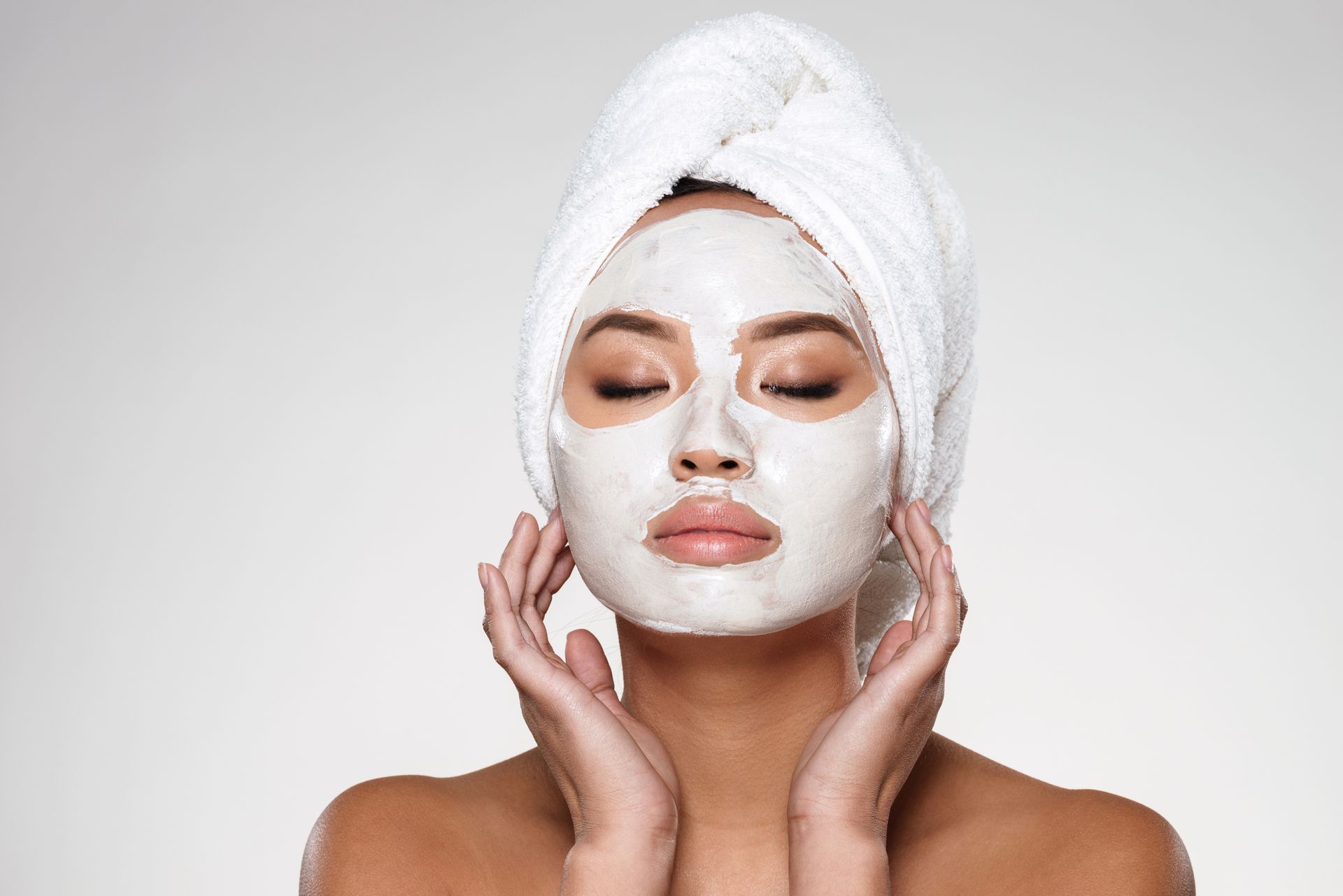 Woman with a white facial mask, eyes closed, hands near cheeks, wearing a towel on her head.