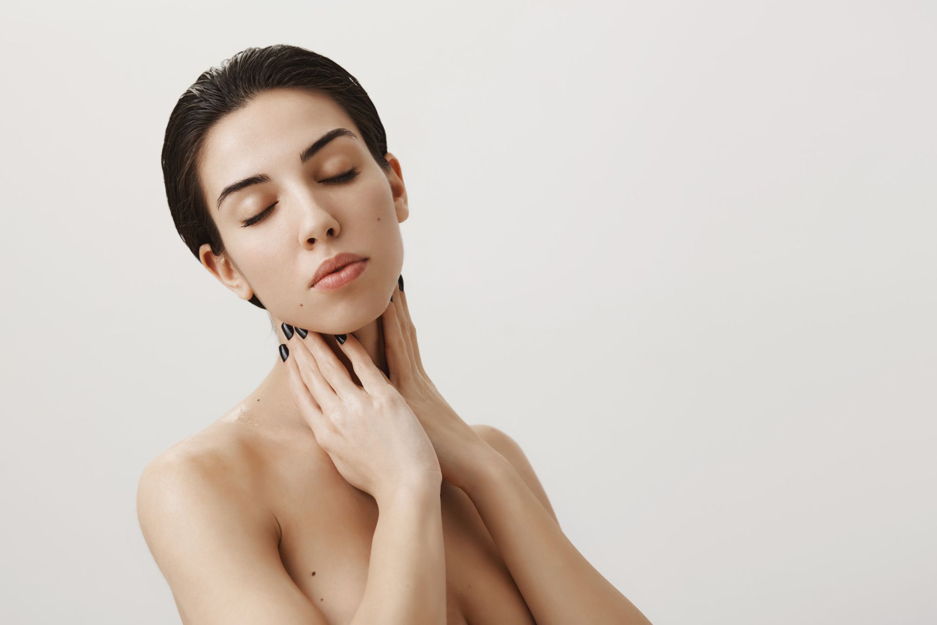 Woman with short dark hair, eyes closed, touching her neck. Neutral background.