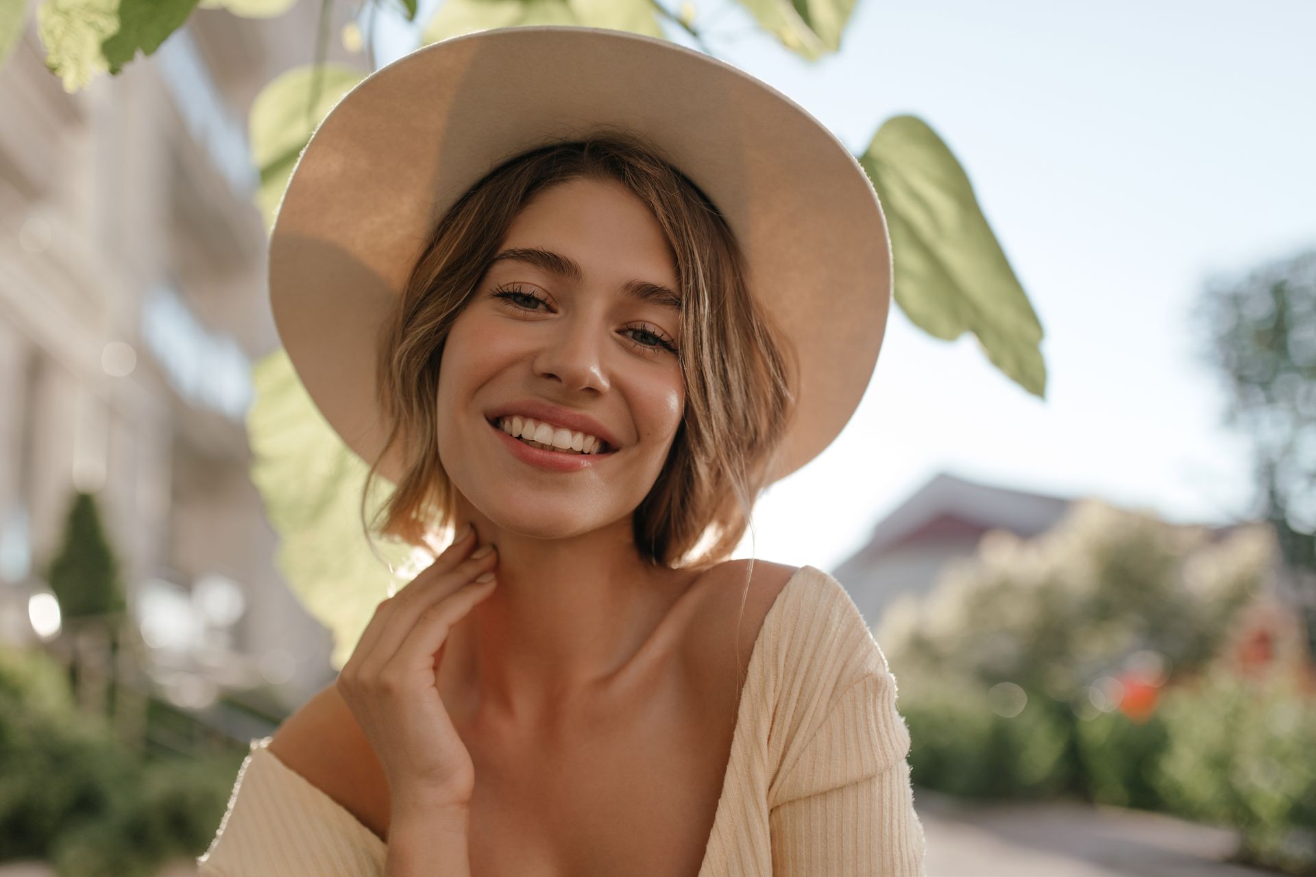 Woman in a beige hat and sweater smiles, touches neck outdoors.