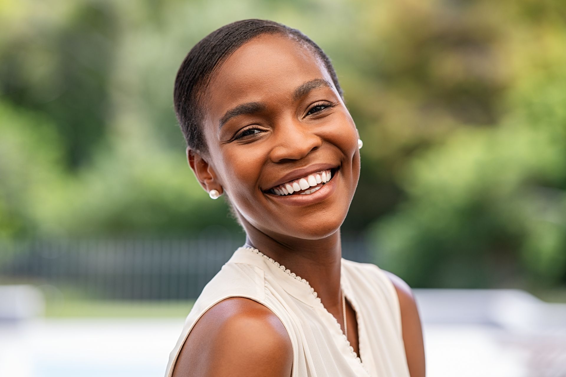 Woman smiling widely outdoors, wearing a white top.
