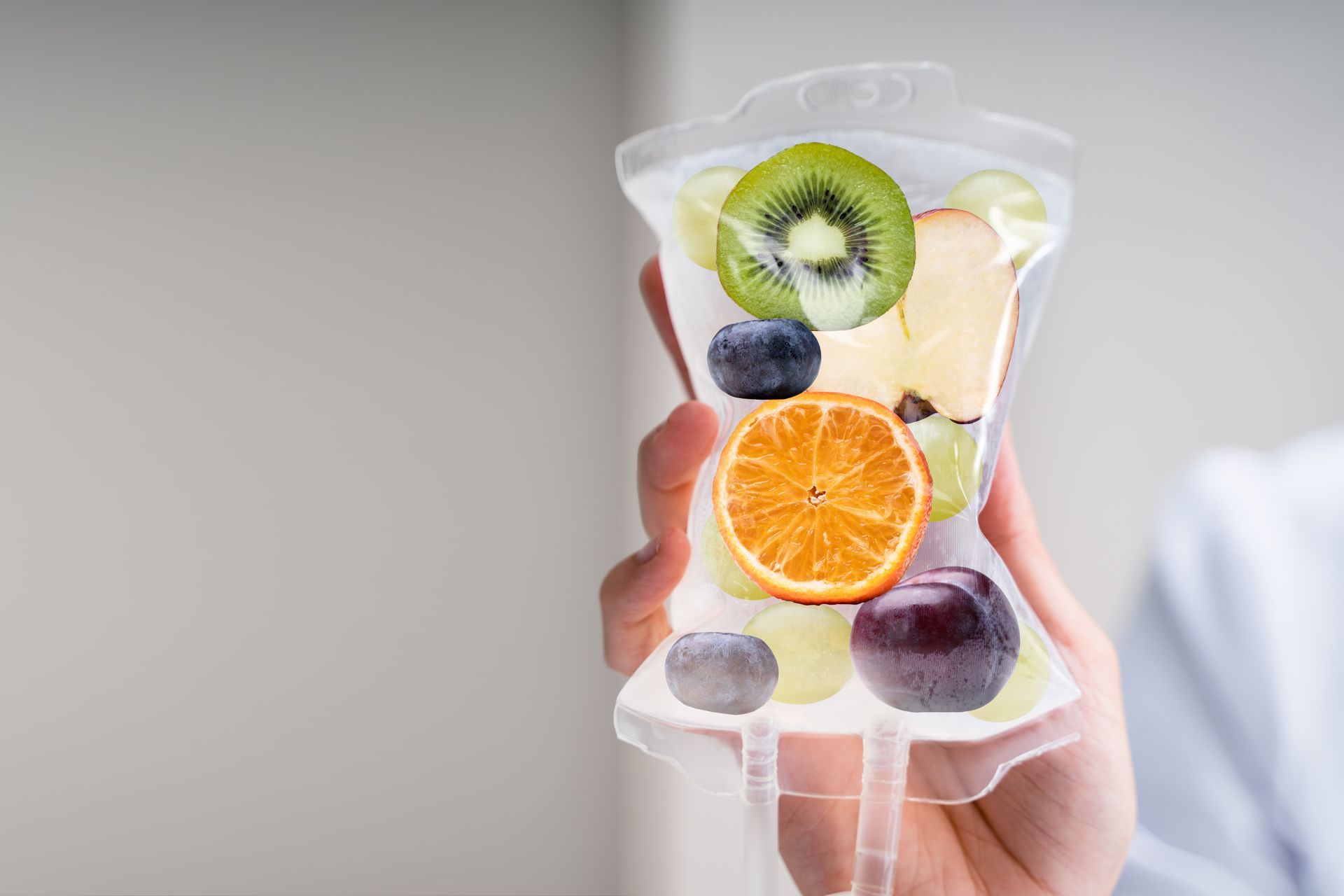 Hand holding an IV bag filled with various fruits like kiwi, orange, and apple.
