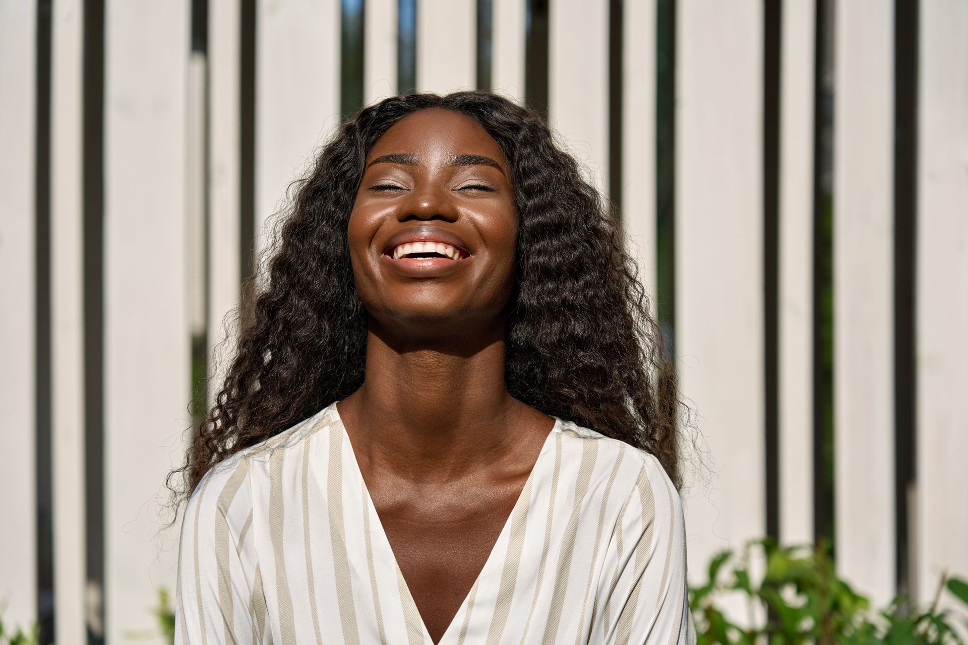Woman with long wavy hair, eyes closed, laughing in front of a white slatted wall, sunny outdoors.