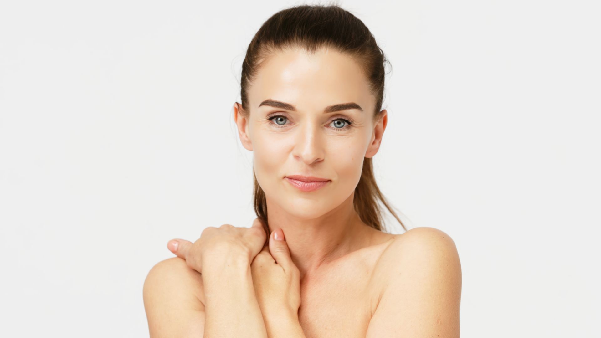 Woman with fair skin and brown ponytail smiles, touching her shoulders, against a white backdrop.