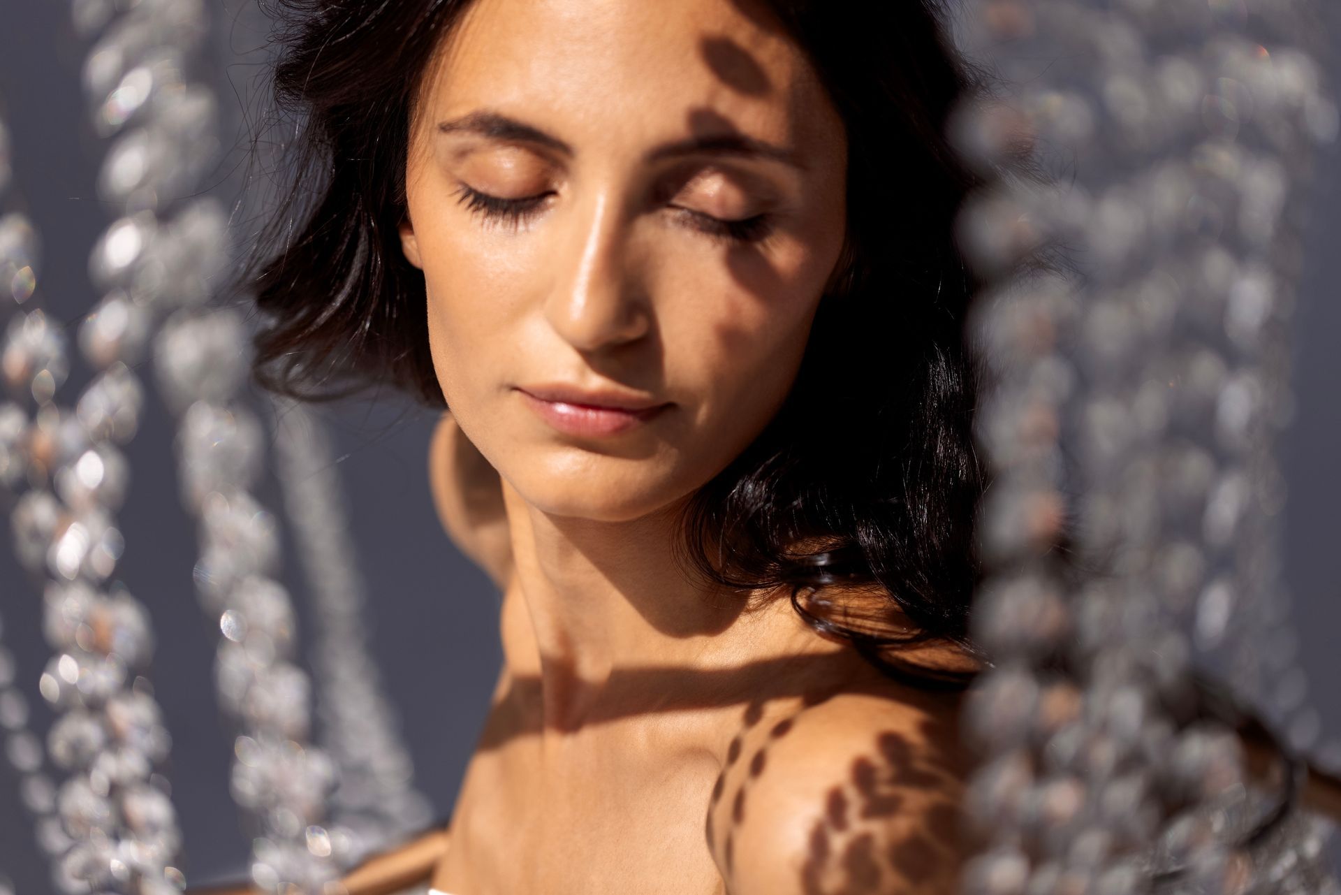 Woman with eyes closed, illuminated by sunlight filtering through beaded strands, casting shadows.