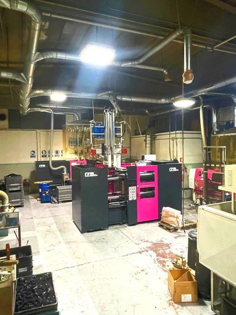 Officina industriale con macchinari neri e rosa, condotti metallici e pavimento ingombro sotto luci intense dall'alto.