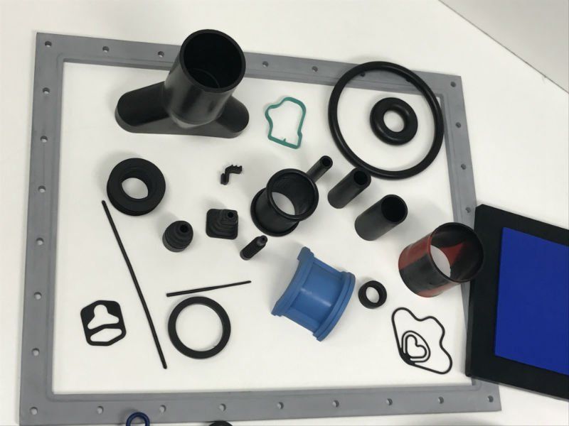 Vari pezzi di gomma e plastica neri e grigi disposti su una superficie bianca, con un pezzo blu e il bordo dello schermo visibili.