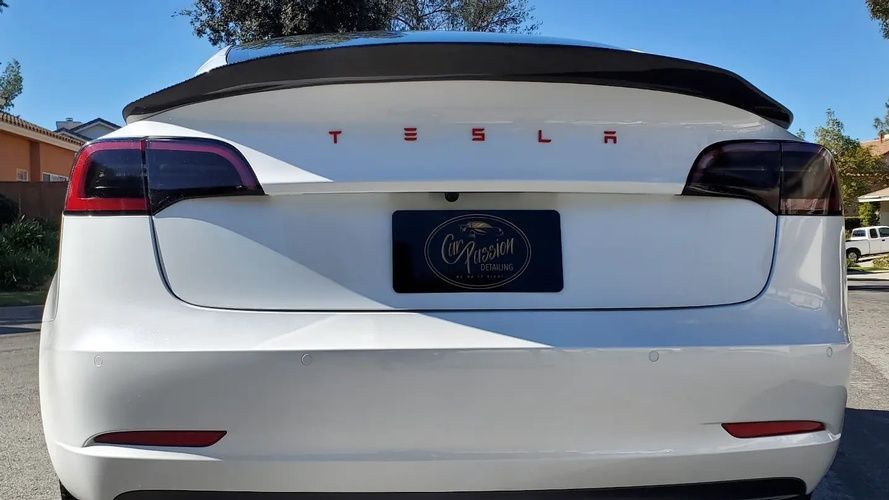 A white tesla model 3 with a black spoiler is parked in a driveway.