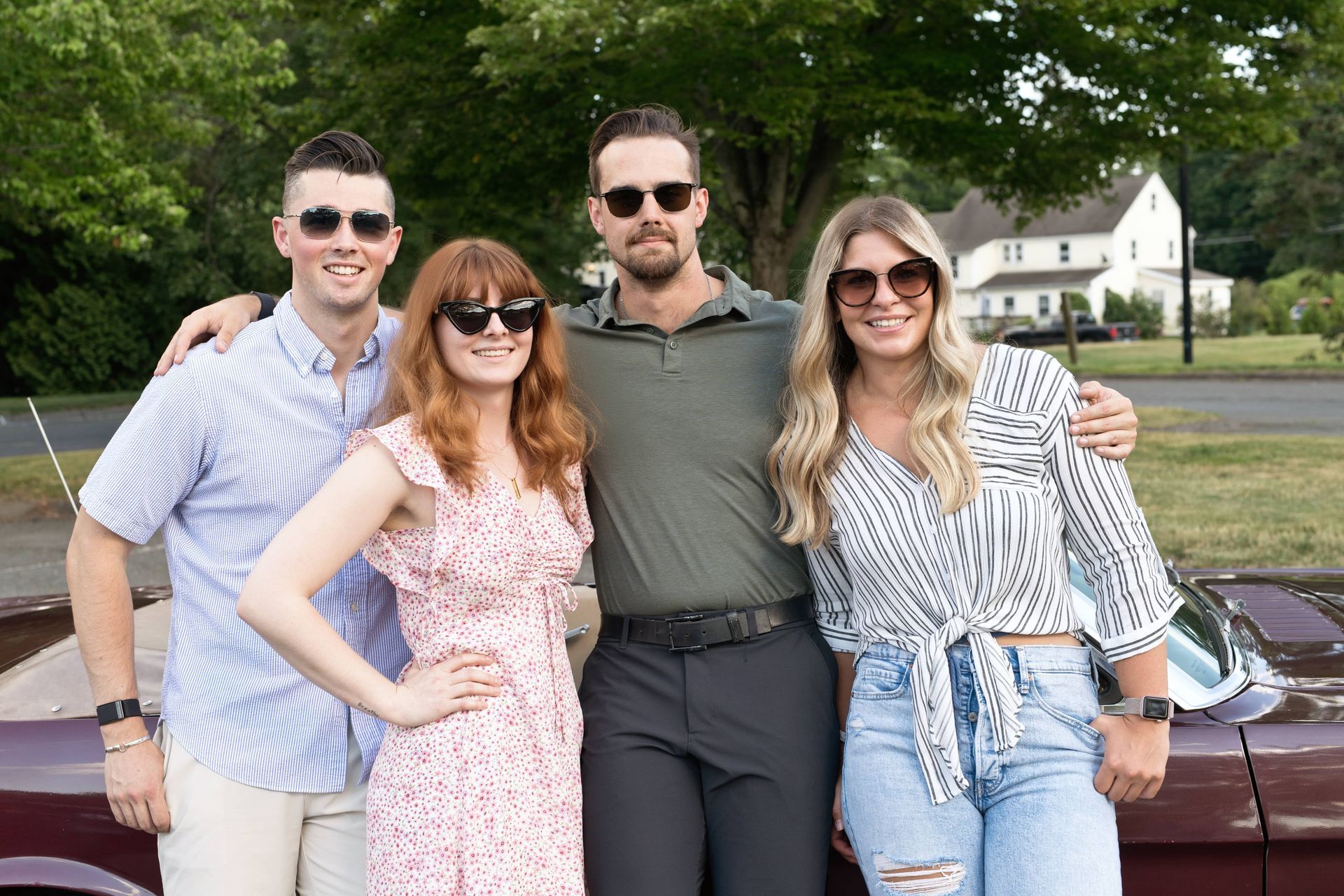 Four friends pose smiling outdoors by a car, with trees and houses in the background.
