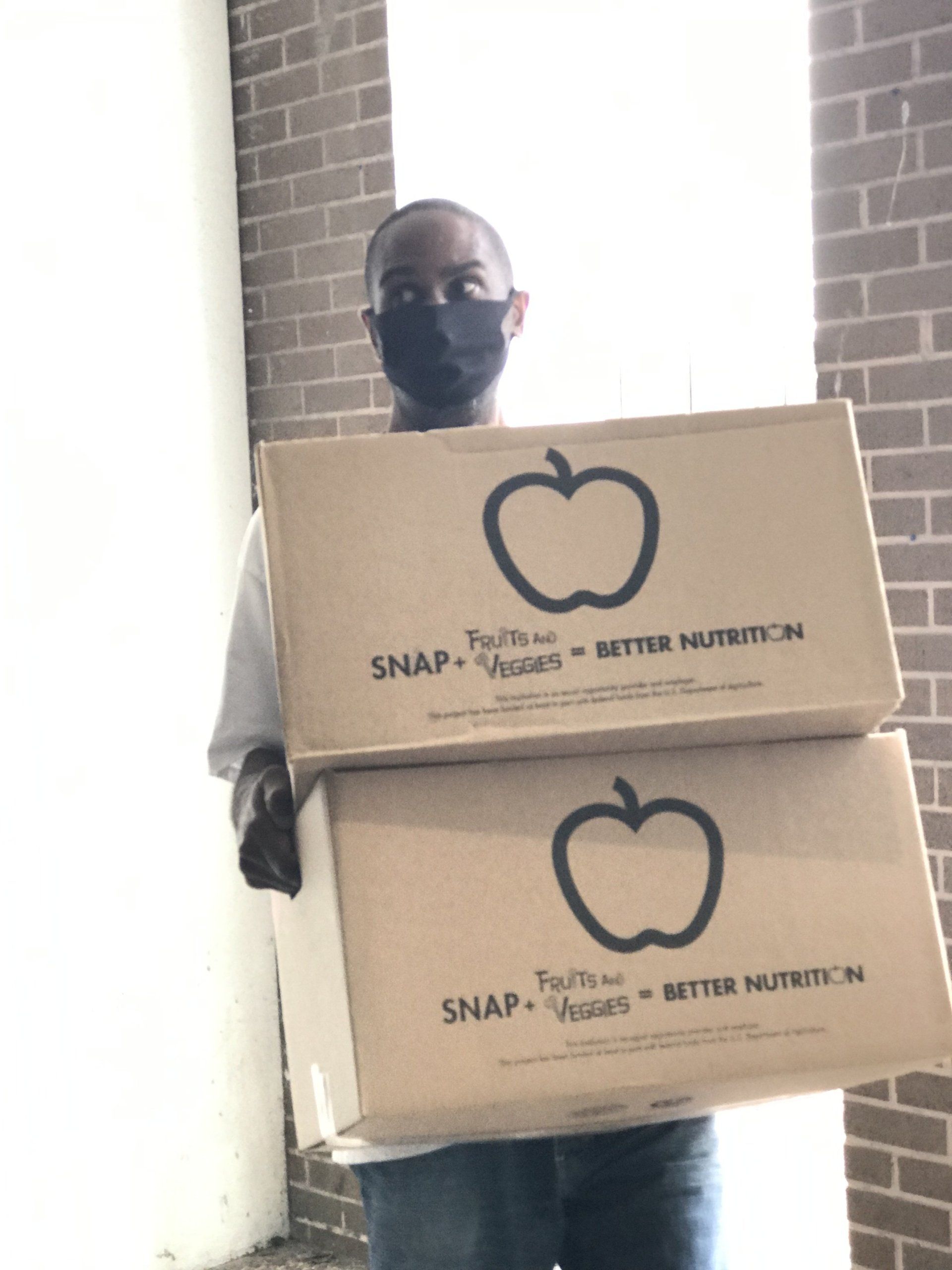 A man wearing a mask is holding two boxes with apples on them.