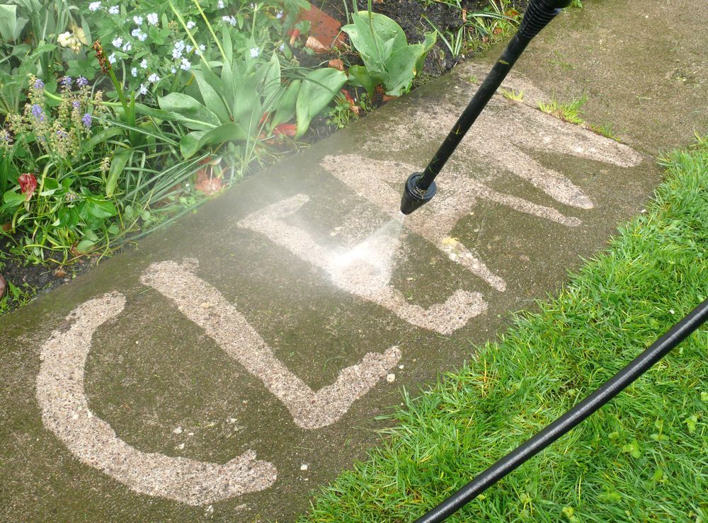 Pressure washer cleaning “LEAP” painted on a concrete path beside garden plants.